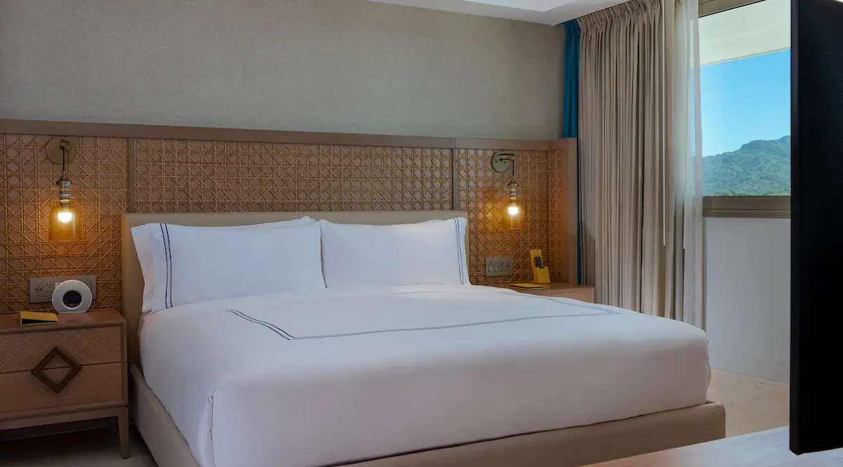 Una moderna habitación de hotel con una cama grande y bien hecha, mesitas de noche, lámparas y una ventana con vista a la montaña.