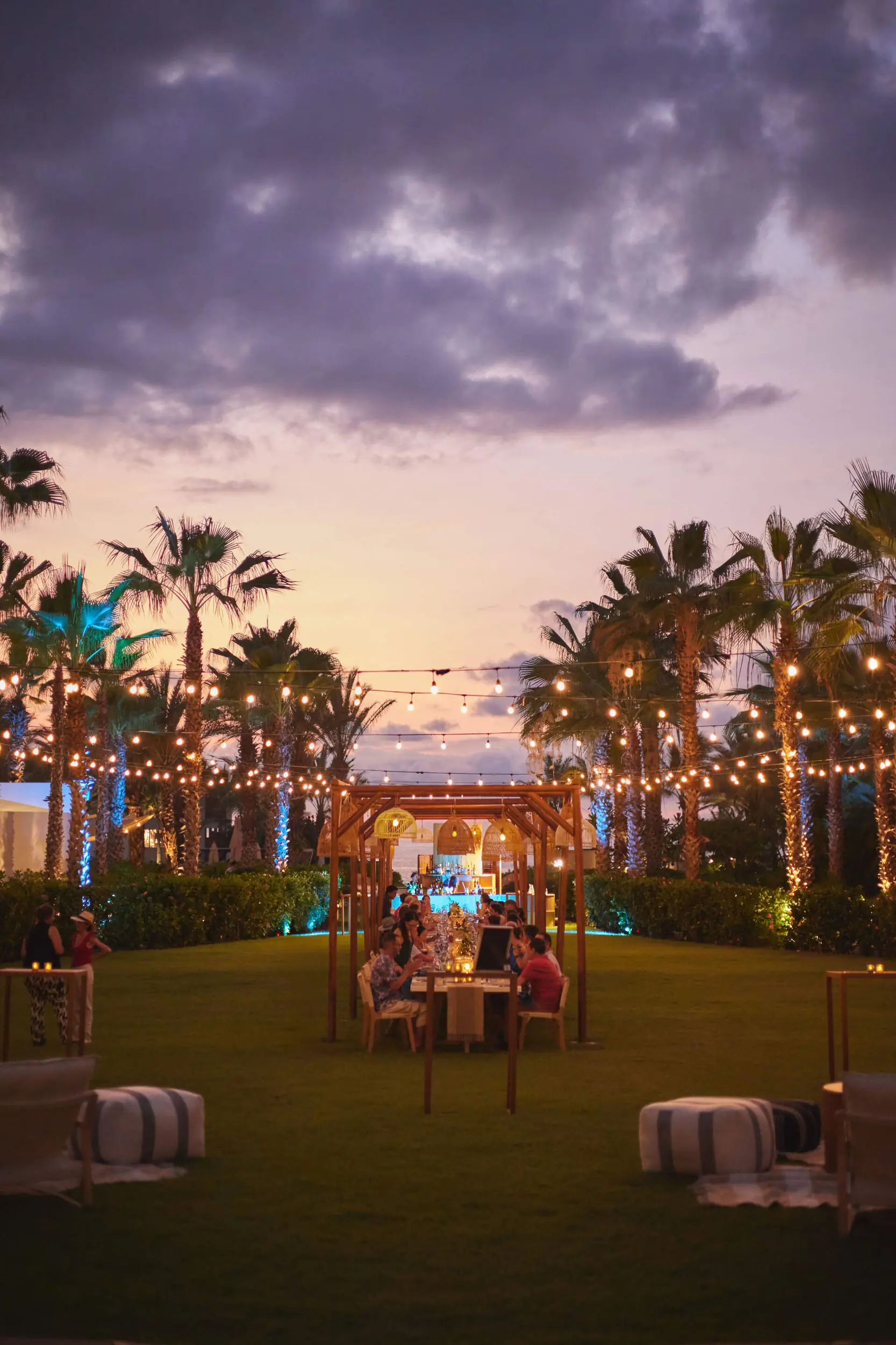 Dinner guests sit under string lights and palm trees, enjoying an elegant outdoor evening event at sunset.