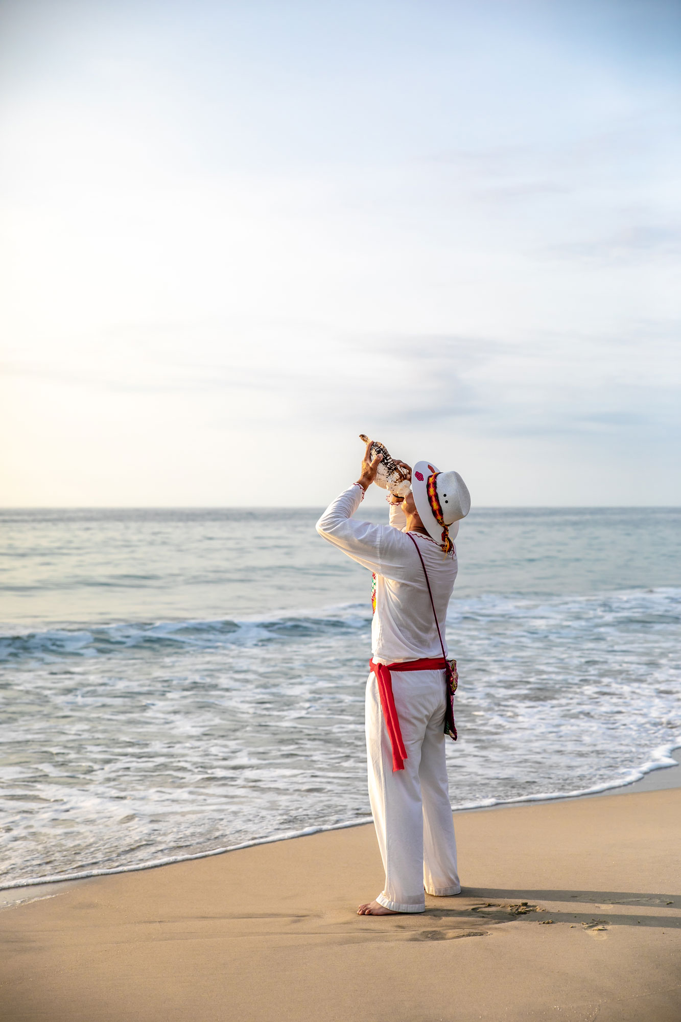 A man in white attire honks a conch shell at the seaside.