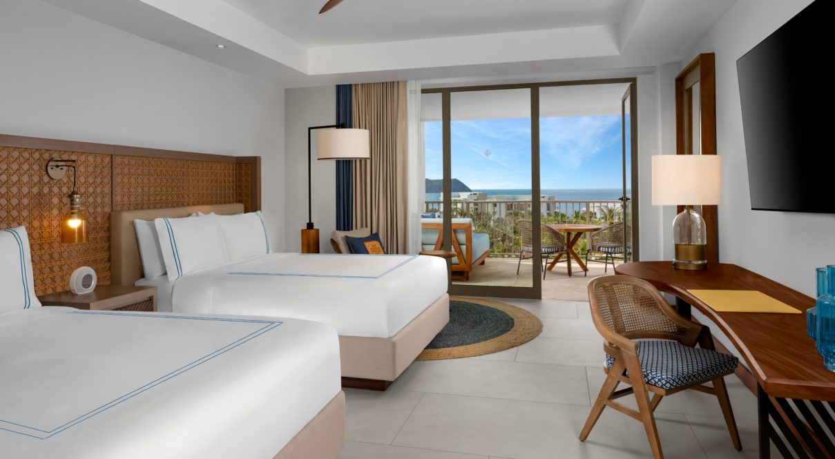 Una habitación de hotel con dos camas, lámparas, tv y silla con vista al río con balcón.