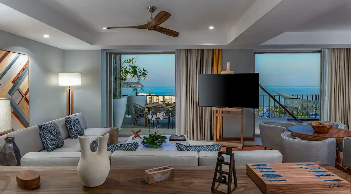 Una sala de estar con sofá, lámpara, tv y cuadro con vista al río con balcón.
