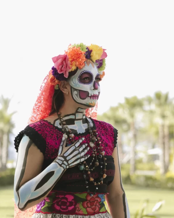 Persona con corona de flores colorida, vestido tradicional bordado, pintura corporal de esqueleto y collares se encuentra al aire libre.