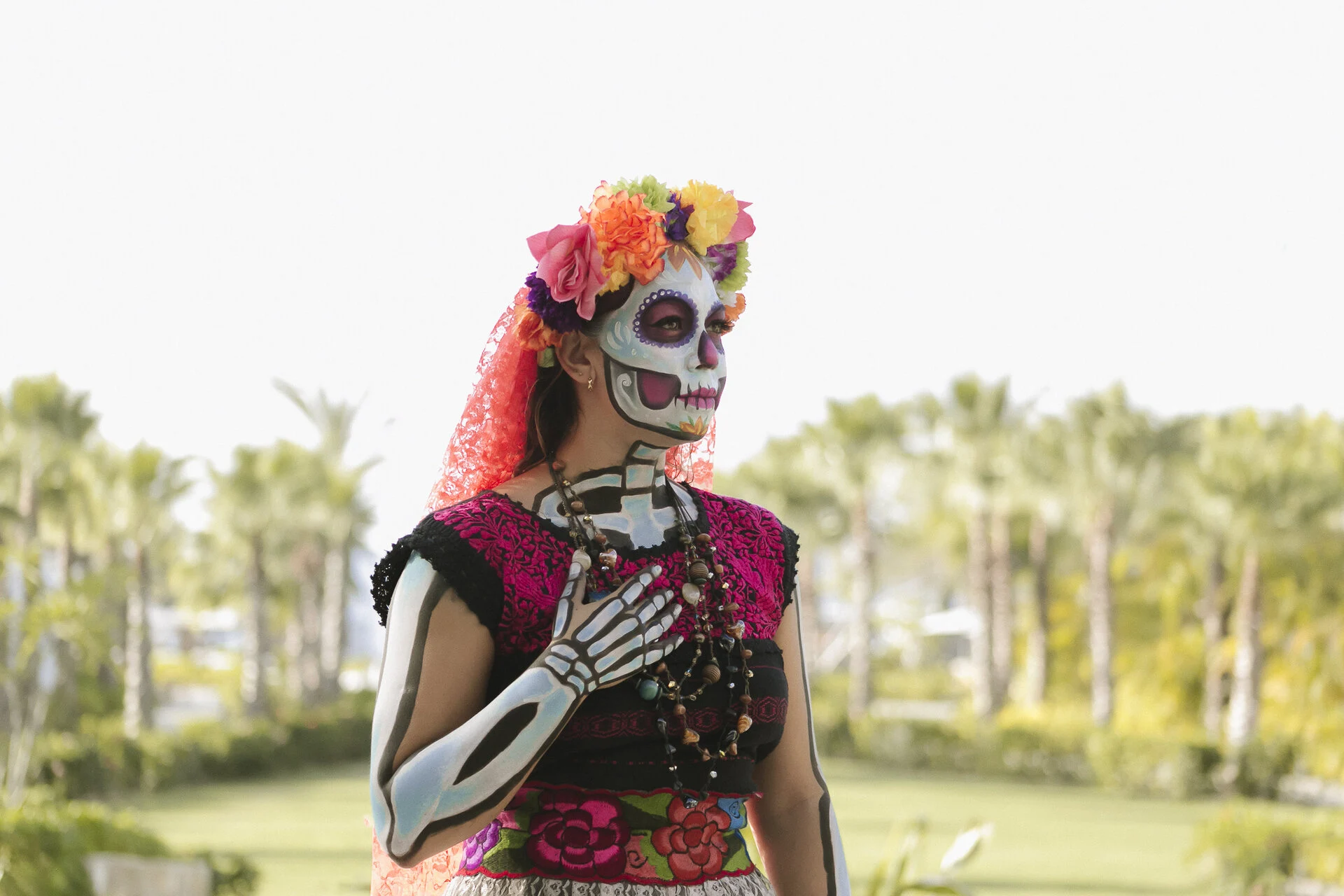 Persona con corona de flores colorida, vestido tradicional bordado, pintura corporal de esqueleto y collares se encuentra al aire libre.