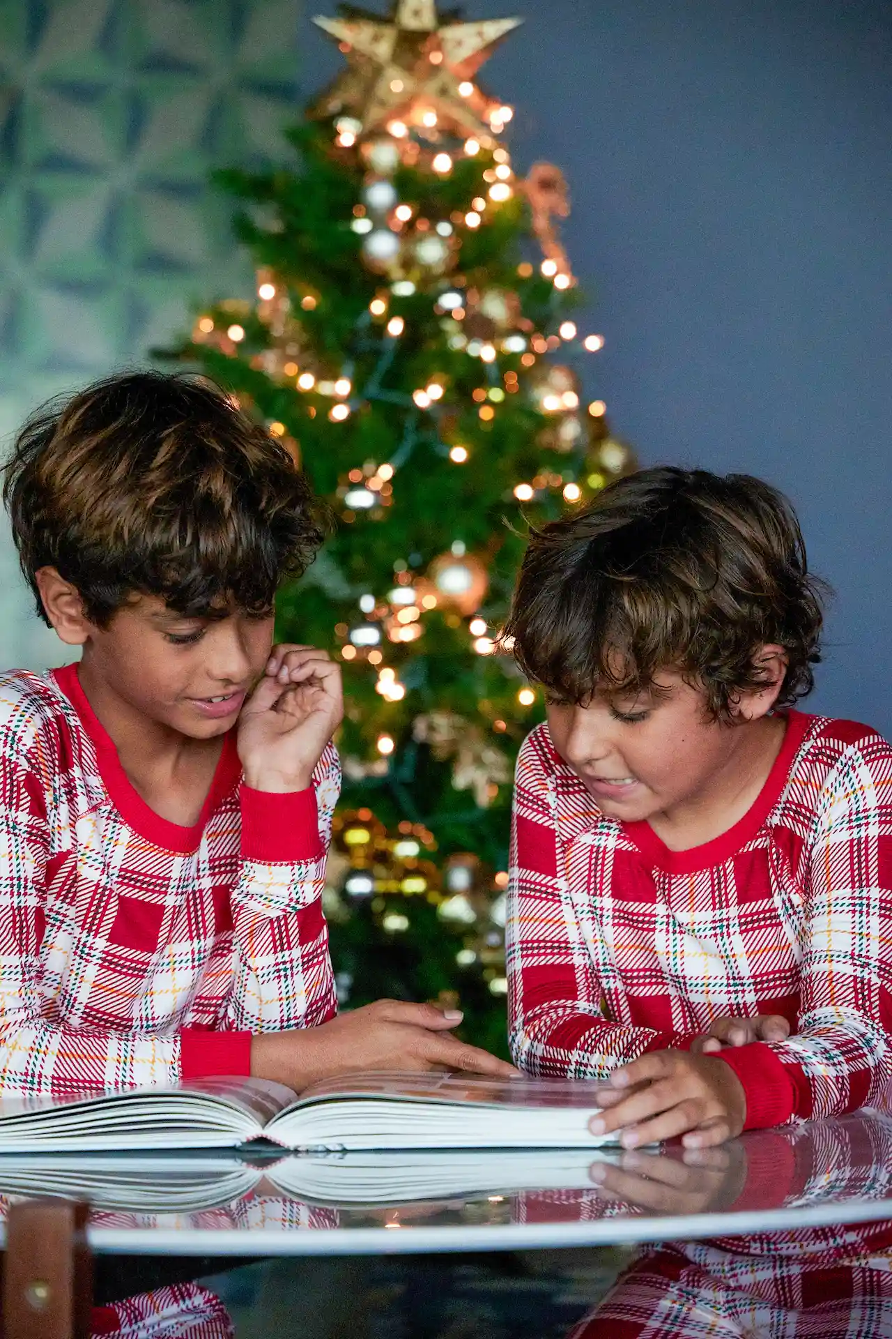 Two kids dressed as Santa read a book at the table by the Christmas tree.