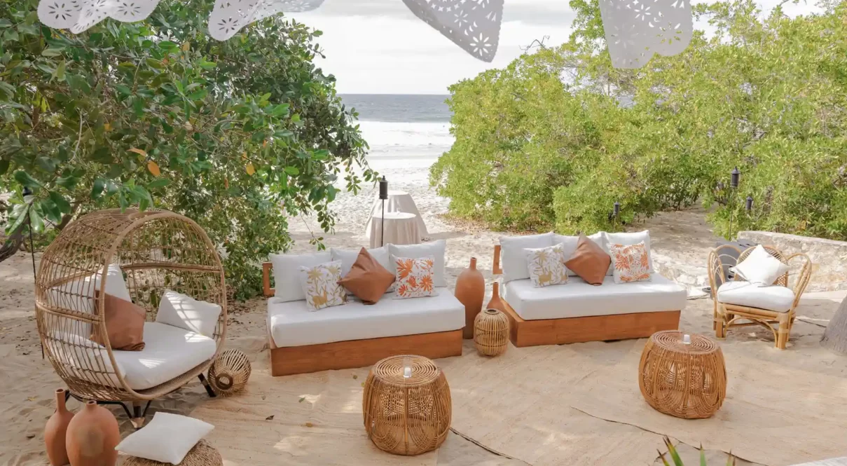 Beachside lounge area with wicker furniture, decorative pillows, clay vases, and festive banners facing the ocean.