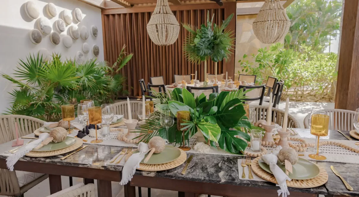 Table setting features tropical leaves, gold glassware, woven chargers, candles, and neutral decor in garden retreat.