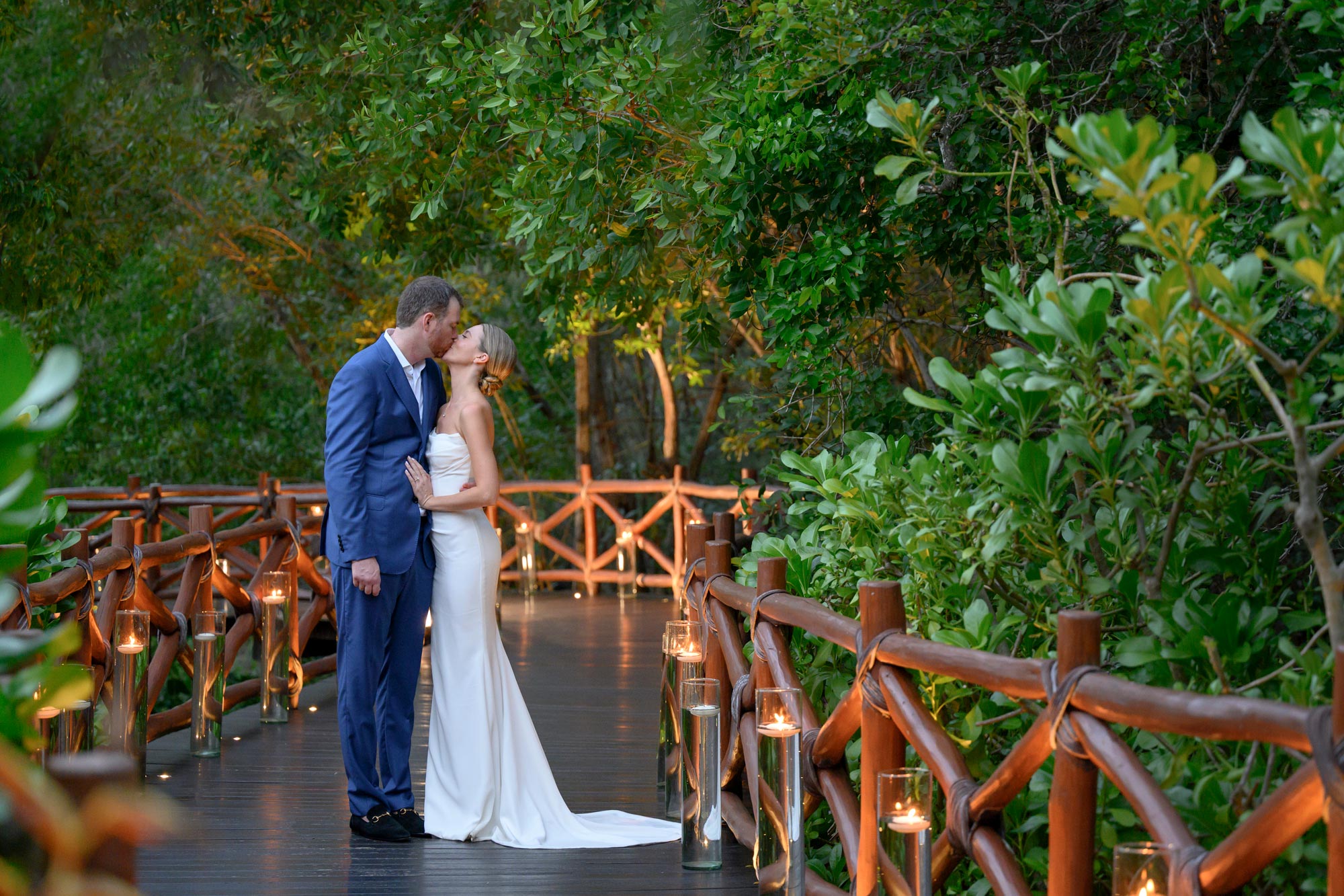 A newlywed couple kisses on a wooden ramp while passing through a dense forest.