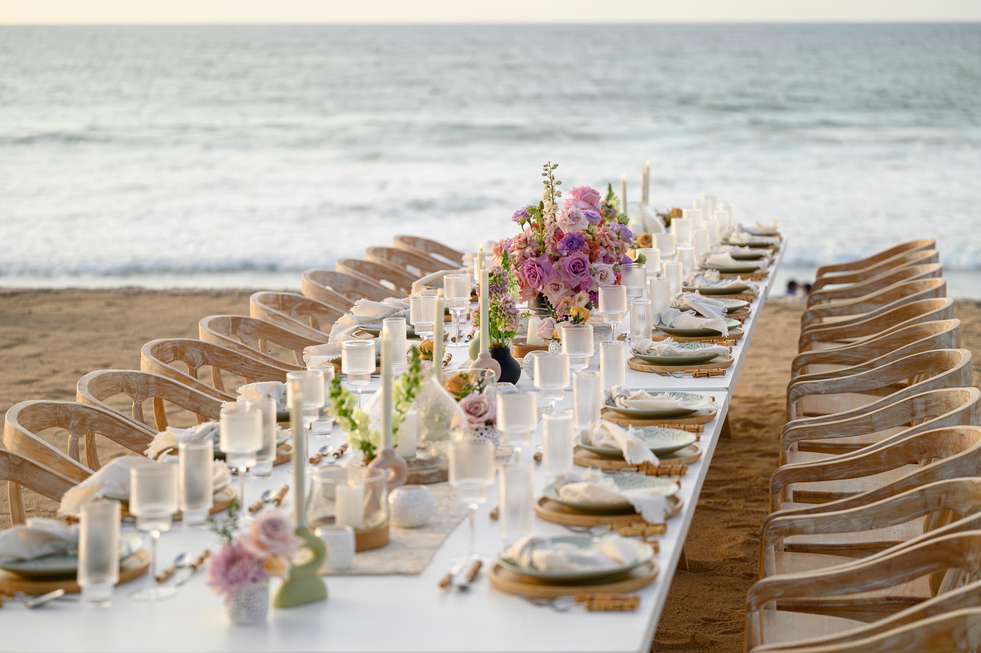 A long dining table is set up on the beach, adorned with flowers and candles.