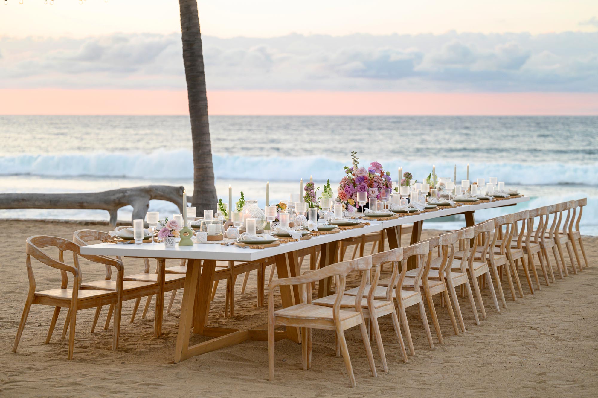 A long dining table is arranged on the beach, decorated with flowers and candles.