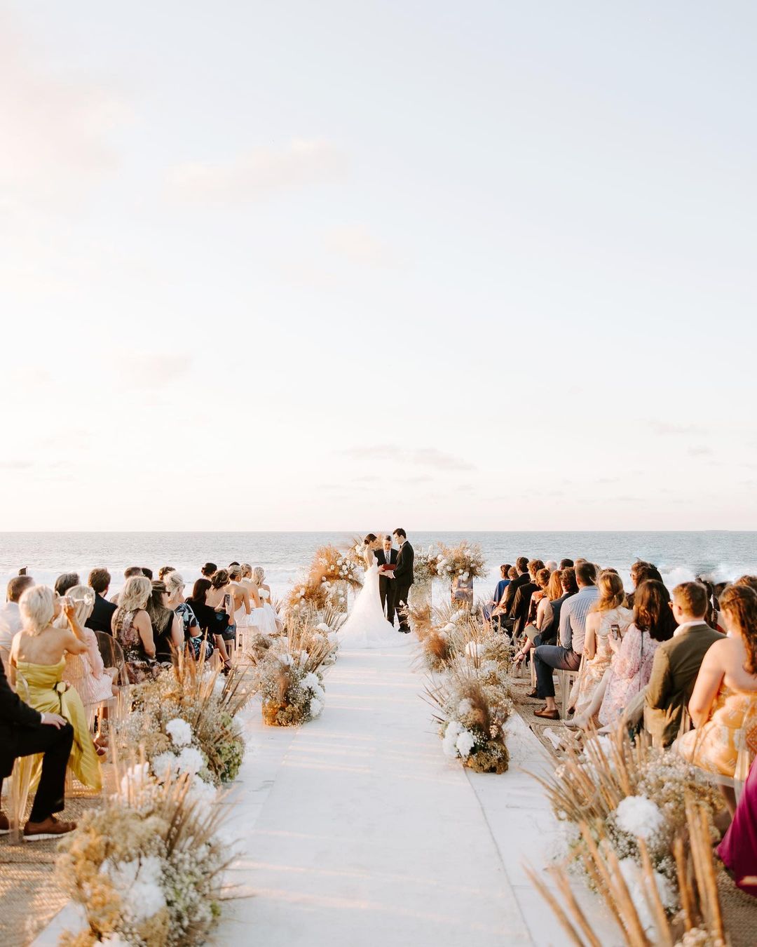 People joyfully witness a ring ceremony seated on either side of a flower-decorated aisle.