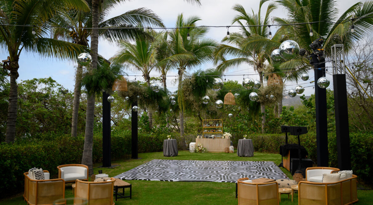 Outdoor lounge area with cane armchairs, patterned dance floor, palm trees, and hanging disco balls overhead.