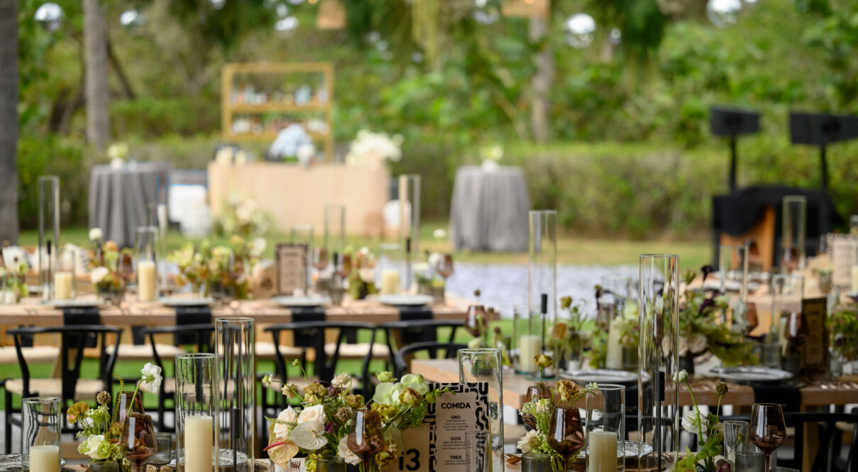 Elegant outdoor event setup with floral arrangements, candles, glassware, and table settings in a lush garden.
