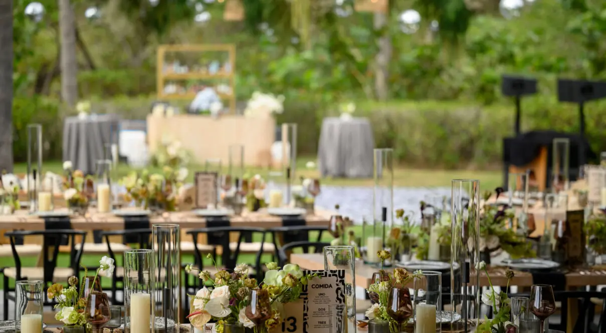 Elegant outdoor event setup with floral arrangements, candles, glassware, and table settings in a lush garden.