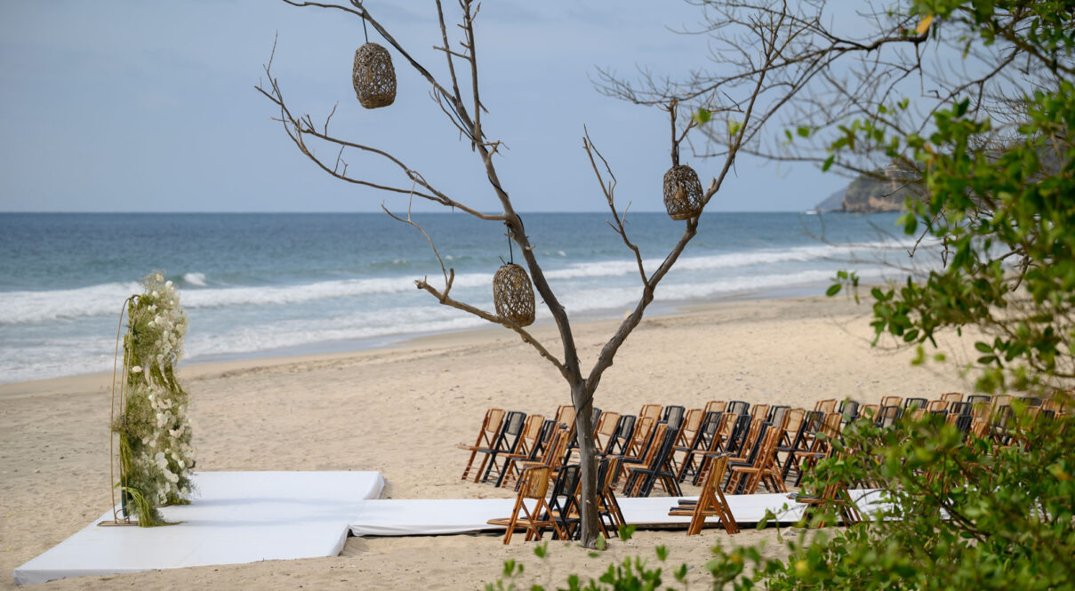 Beach wedding ceremony setup features folding chairs, flower arch, rustic tree with lanterns, white platform, ocean view.