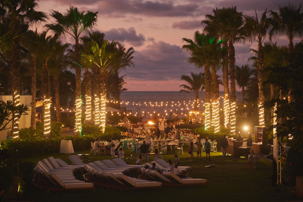 Outdoor evening event with palm trees wrapped in lights, lounge chairs, string lights, and ocean sunset backdrop