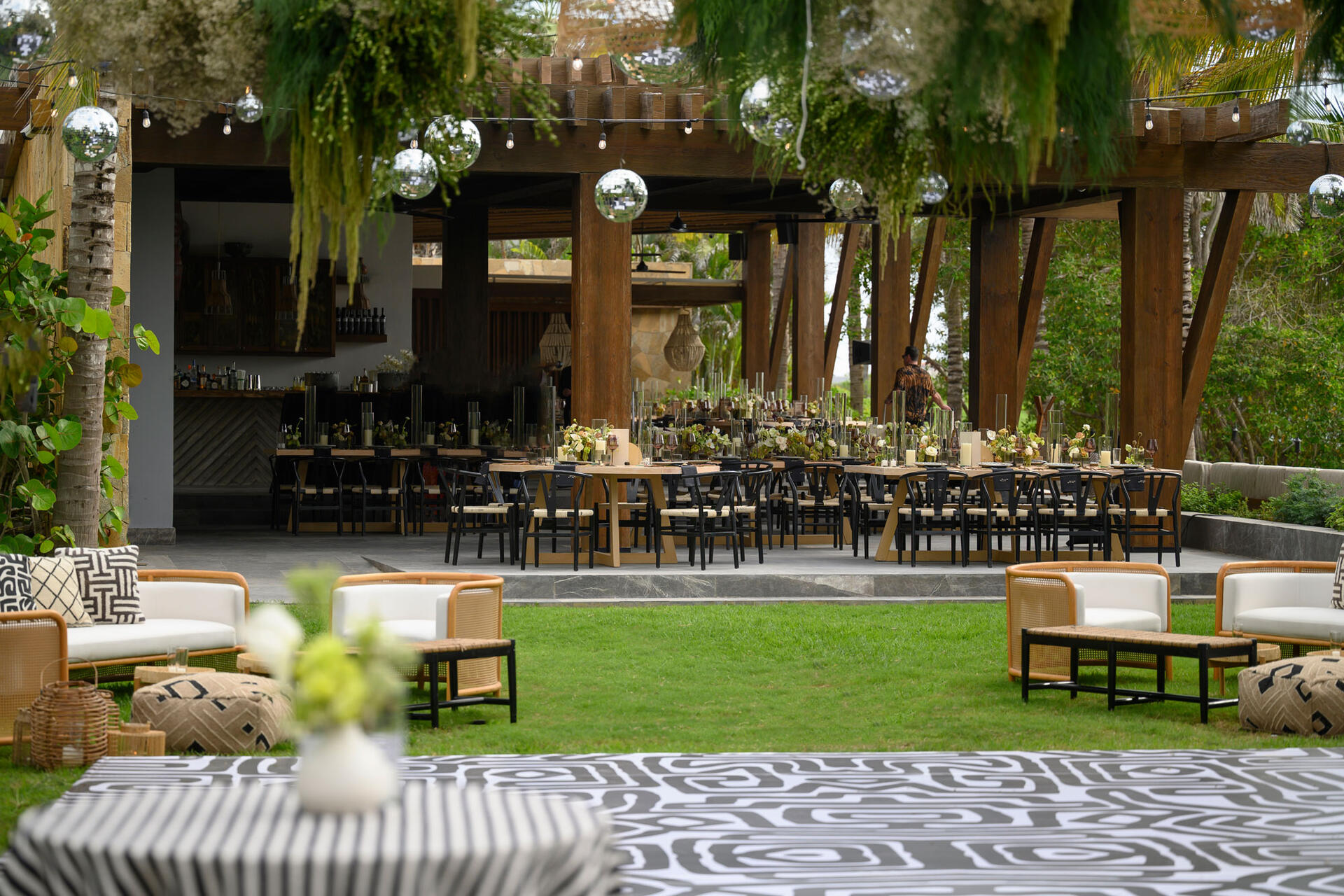 Modern outdoor event setup with black chairs, floral décor, disco balls, patterned tables, and cozy lounge seating.