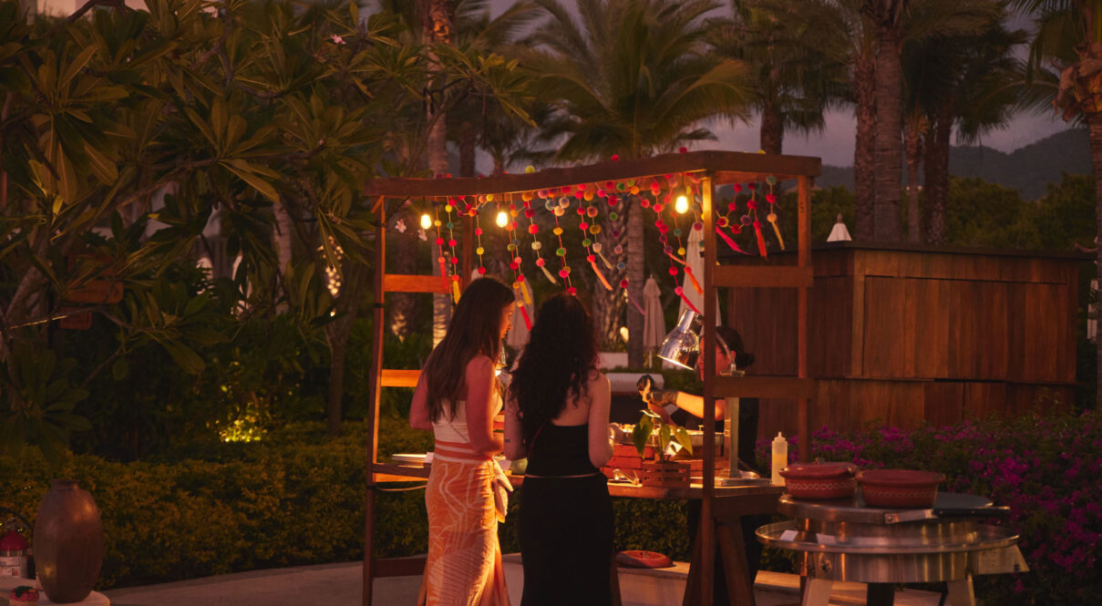 Dos mujeres esperan en un puesto de comida festiva al aire libre, decorado con luces de cadena y guirnaldas de pompones durante la noche.