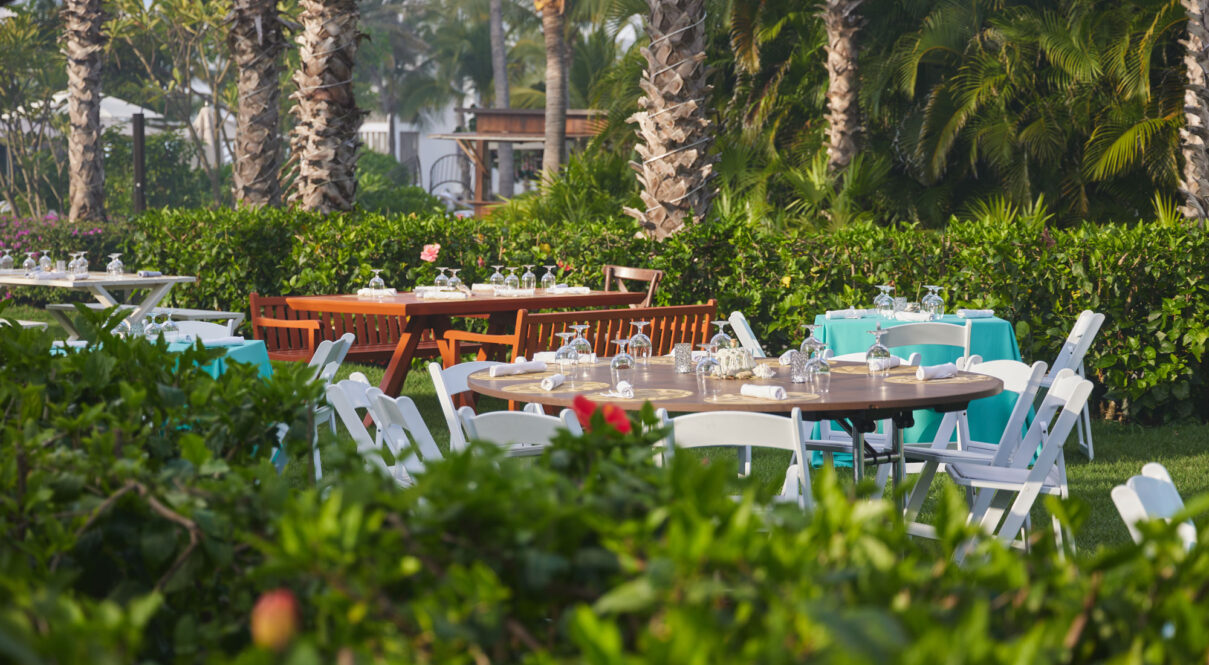 Mesas para eventos redondas y rectangulares con manteles turquesas, sillas blancas y cristalería en un exuberante jardín.