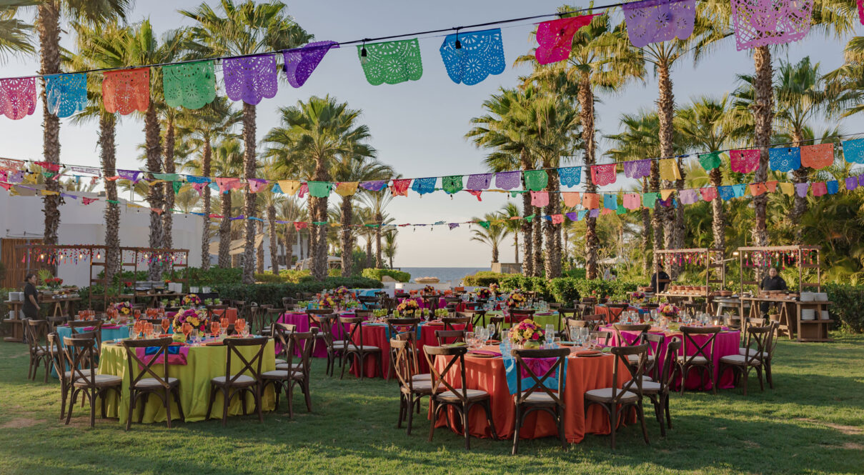 Mesas redondas con manteles de colores, centros de mesa florales, sillas de madera y pancartas de papel picado decoran el jardín festivo.