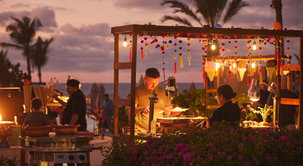 A los invitados se les sirve comida en un puesto de mercado al aire libre decorado con luces festivas y guirnaldas de pompones al atardecer.