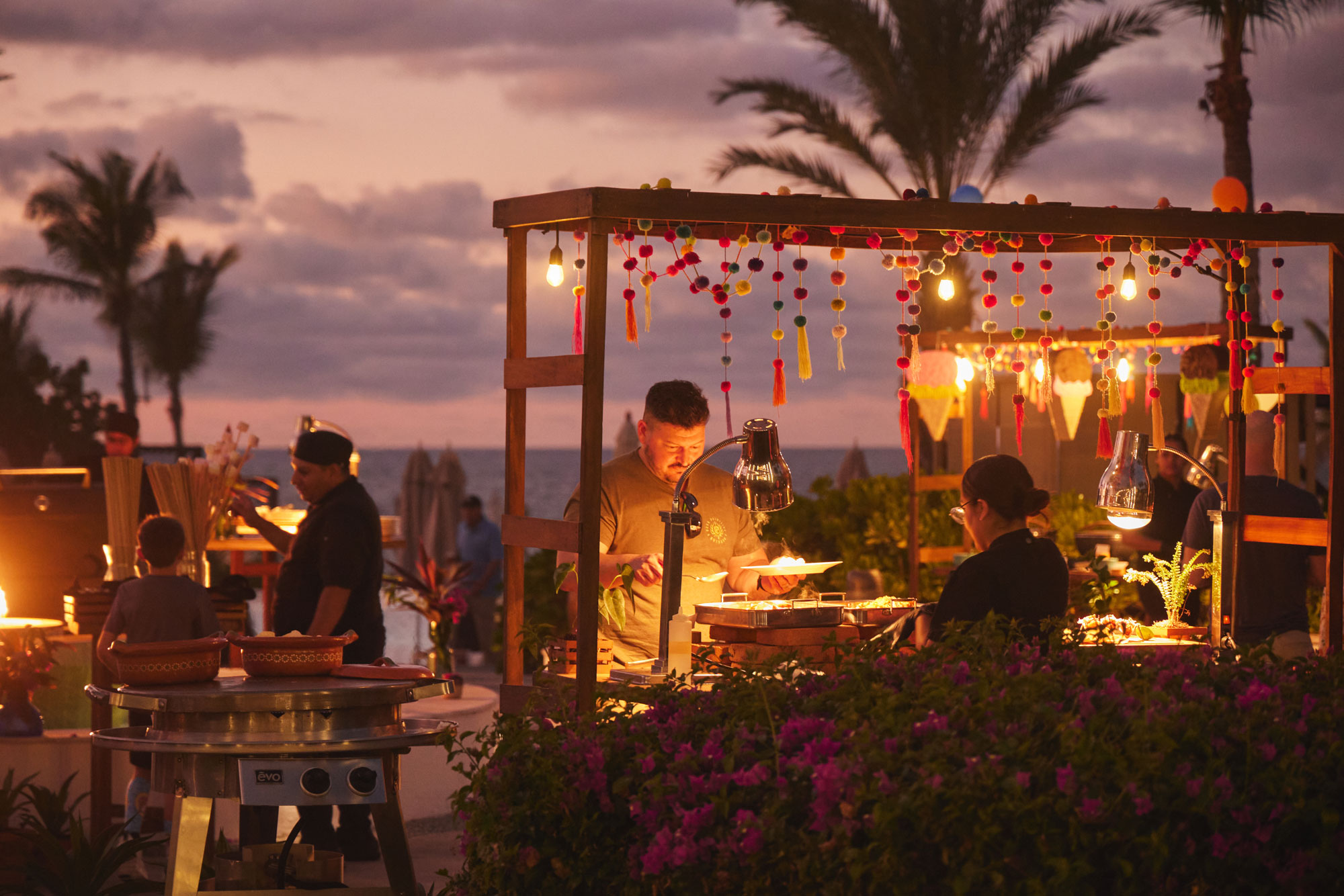Guests being served food at outdoor market stall decorated with festive lights and pompom garlands at sunset.