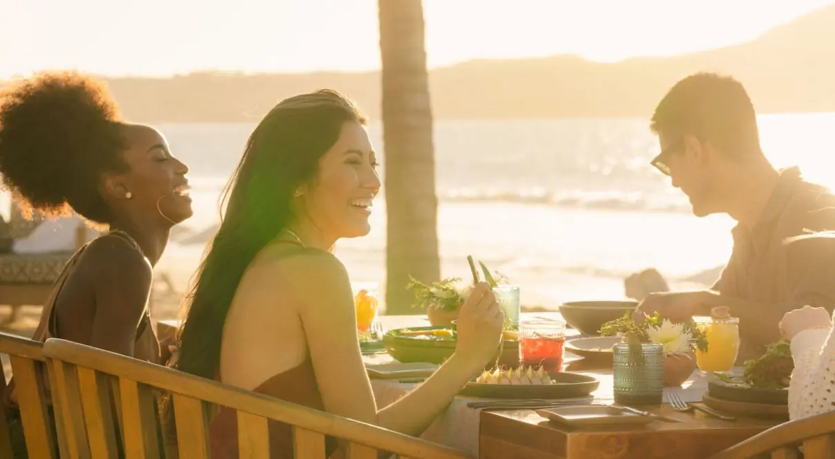 Dos mujeres y un hombre saborean el desayuno junto al mar bajo el sol de la mañana.