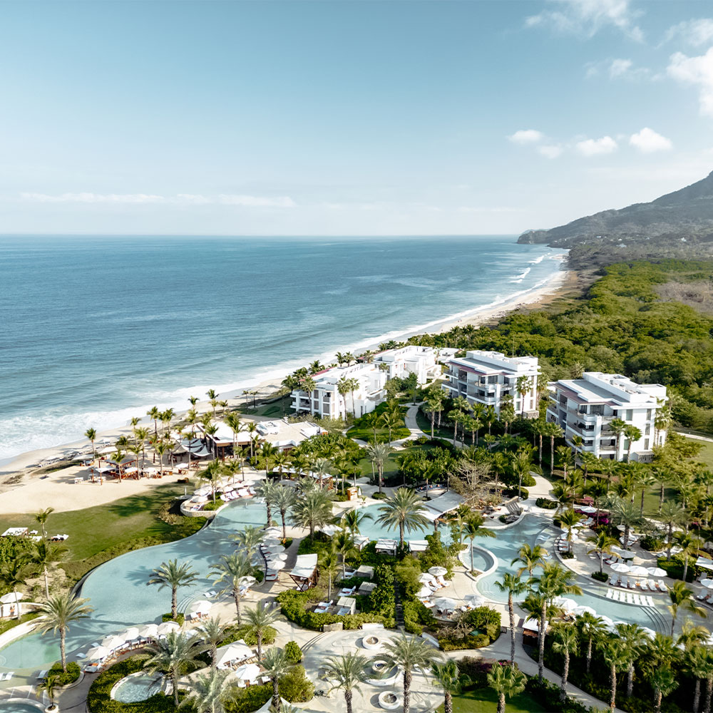 Vista aérea de un lujoso resort frente al mar con piscinas, jardines y edificios blancos junto al océano y las montañas.