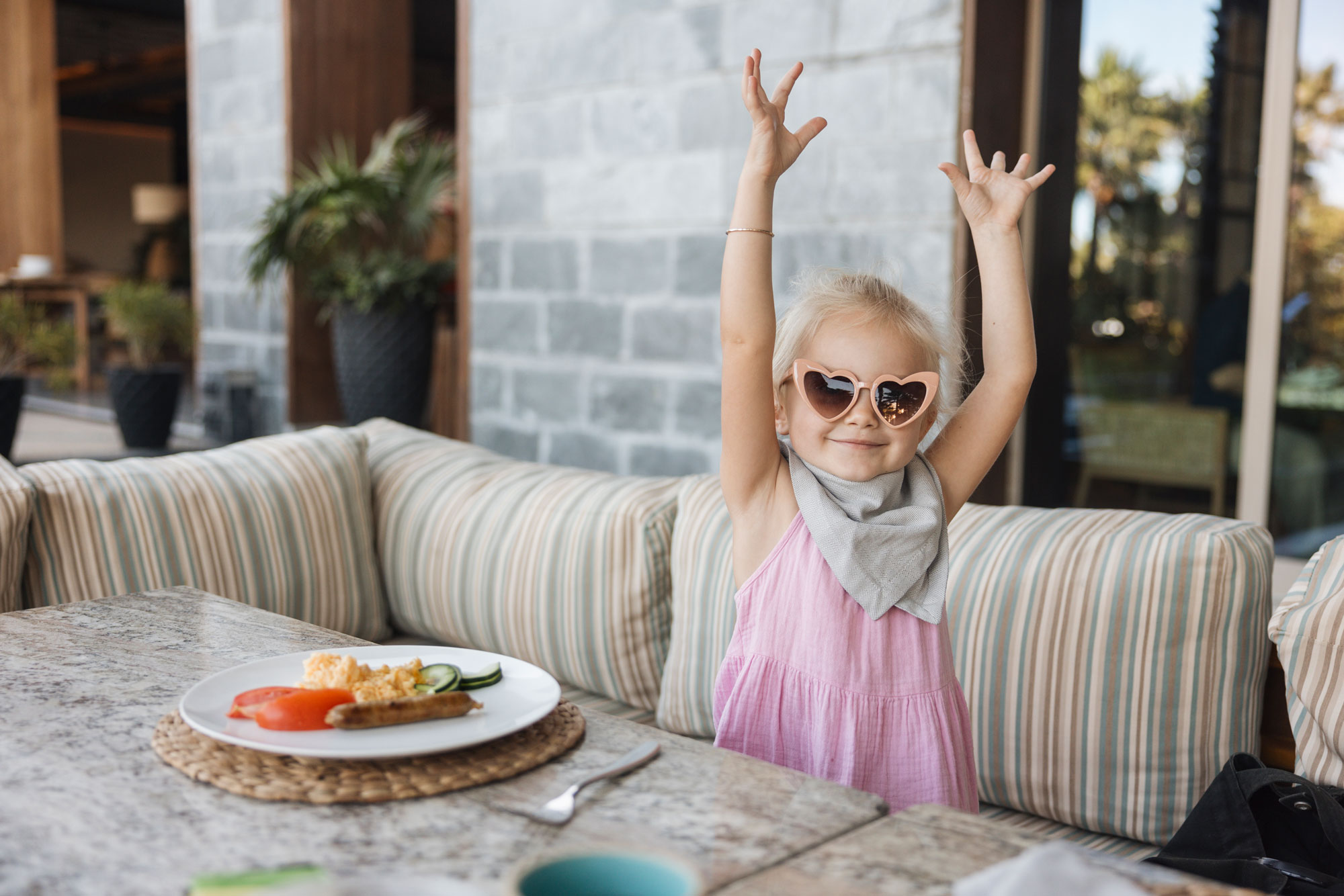 Una joven con un vestido rosa y gafas de sol con forma de corazón levanta los brazos alegremente mientras está sentada al aire libre.