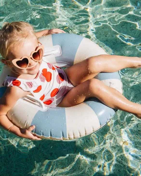Una joven que lleva gafas de sol con forma de corazón se relaja en un flotador de piscina, sonriendo bajo la brillante luz del sol.