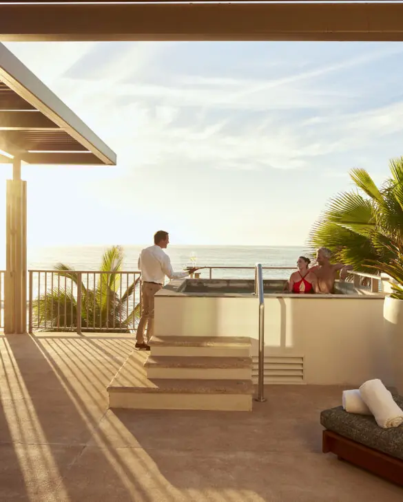 Couple relaxing in rooftop hot tub at sunset while server brings champagne, overlooking ocean.