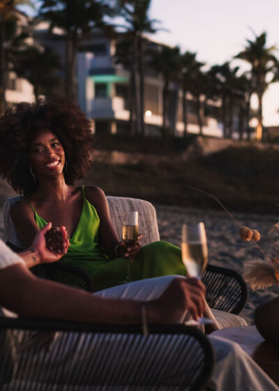 A beach couple on a couch, hands entwined, savoring drinks by the shore.