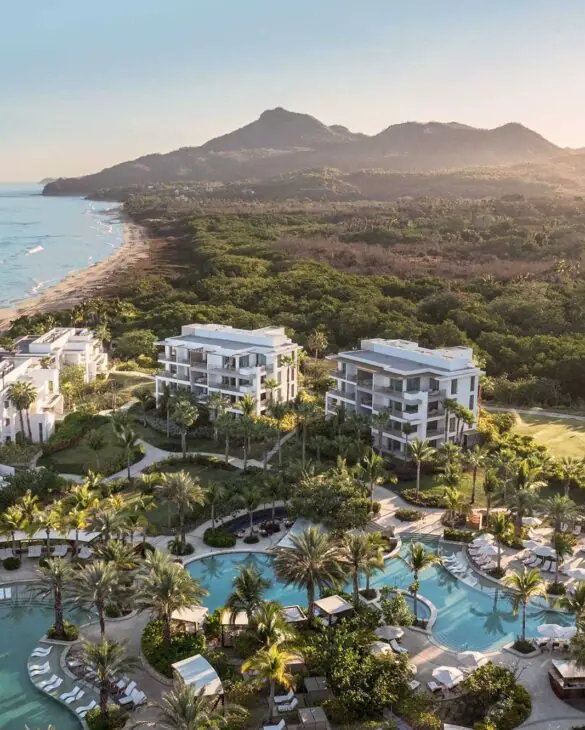 Luxury beachfront resort with white buildings, winding pool, palm trees, and sunset-lit mountains in the background.