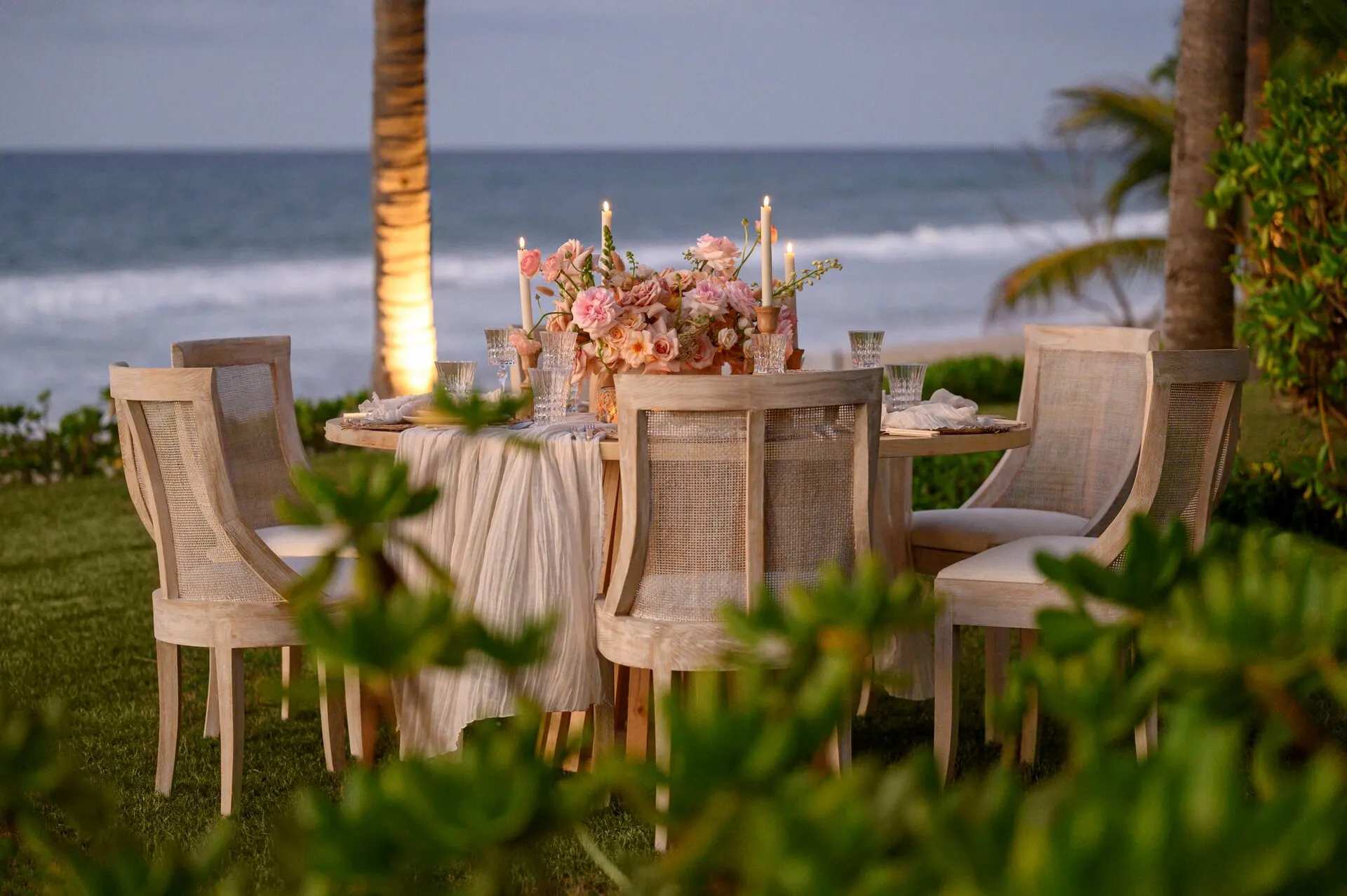 Romantic outdoor table by the ocean with flowers, candles, and wooden chairs.