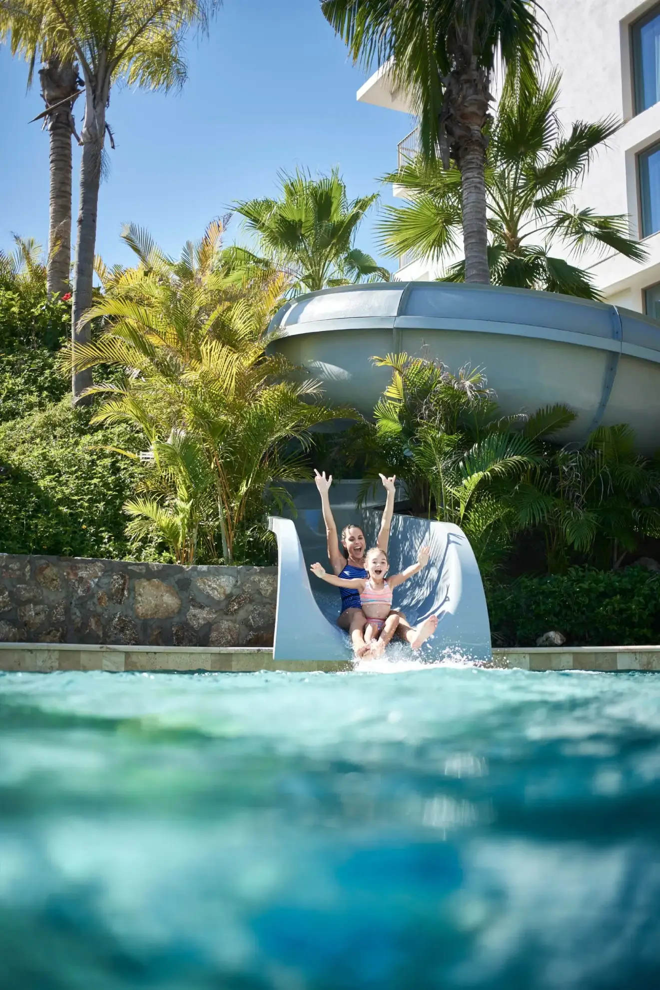 Two children in swimsuits slide down a water slide into a pool, surrounded by palm trees.