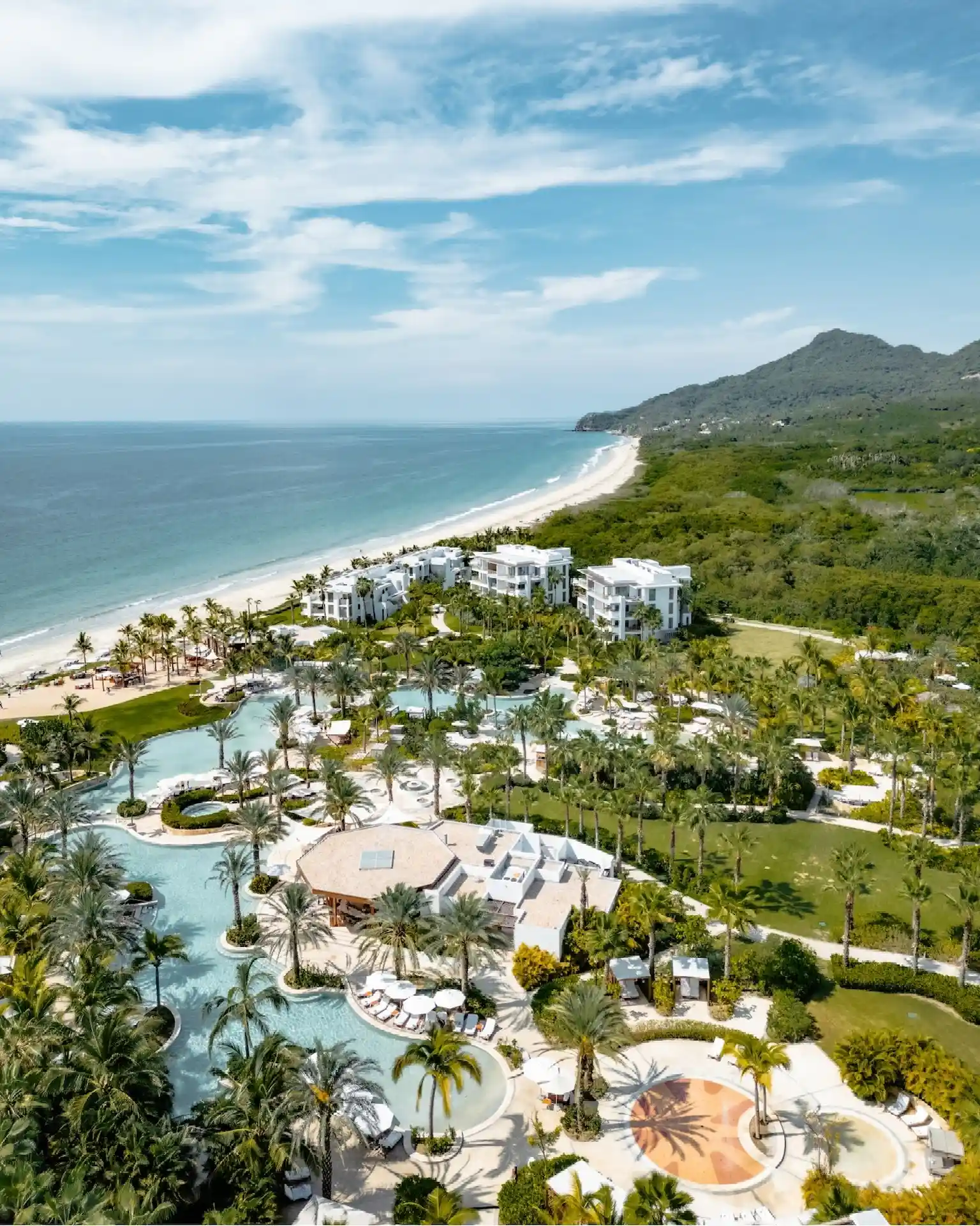 Luxury beachfront resort with pools, palm trees, and ocean views backed by lush green mountains.