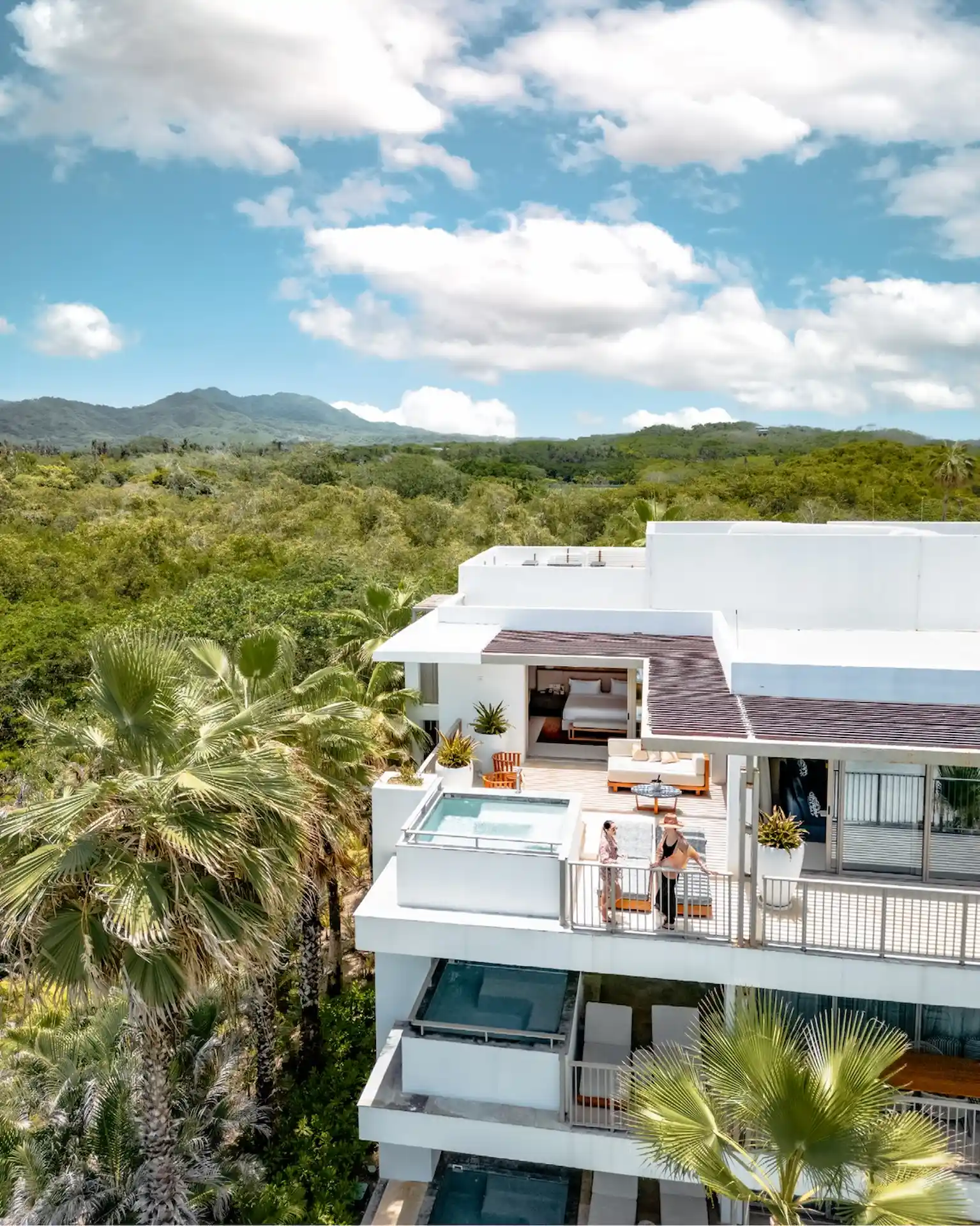 Modern white building with terraces and pools amid tropical greenery and mountain views.