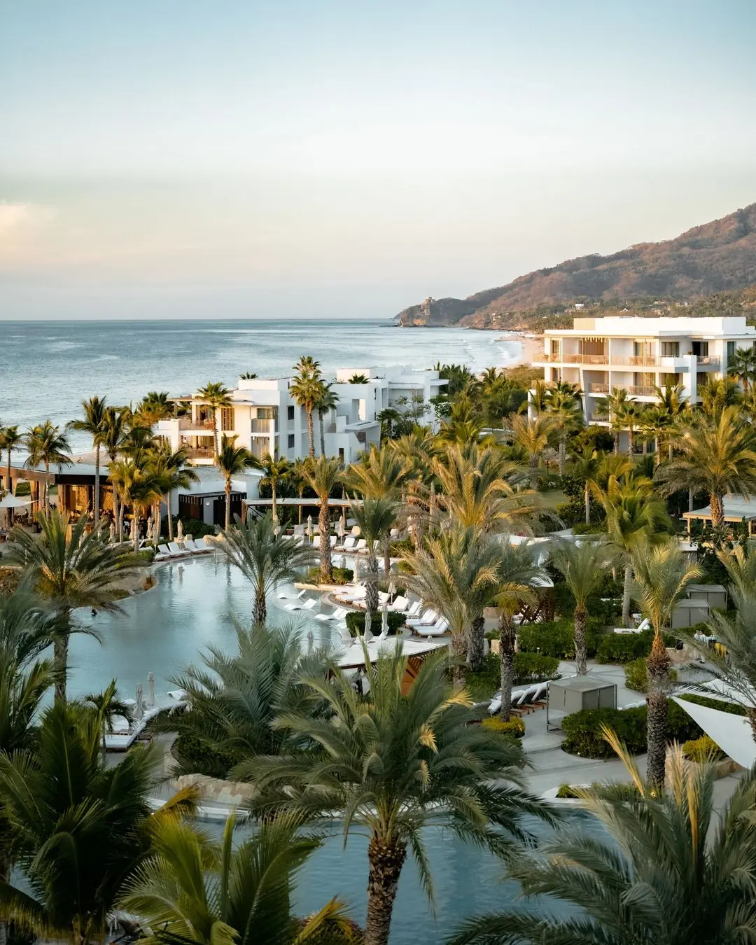 Resort de lujo frente al mar con palmeras, piscinas y modernos edificios blancos con vista a un océano tranquilo.