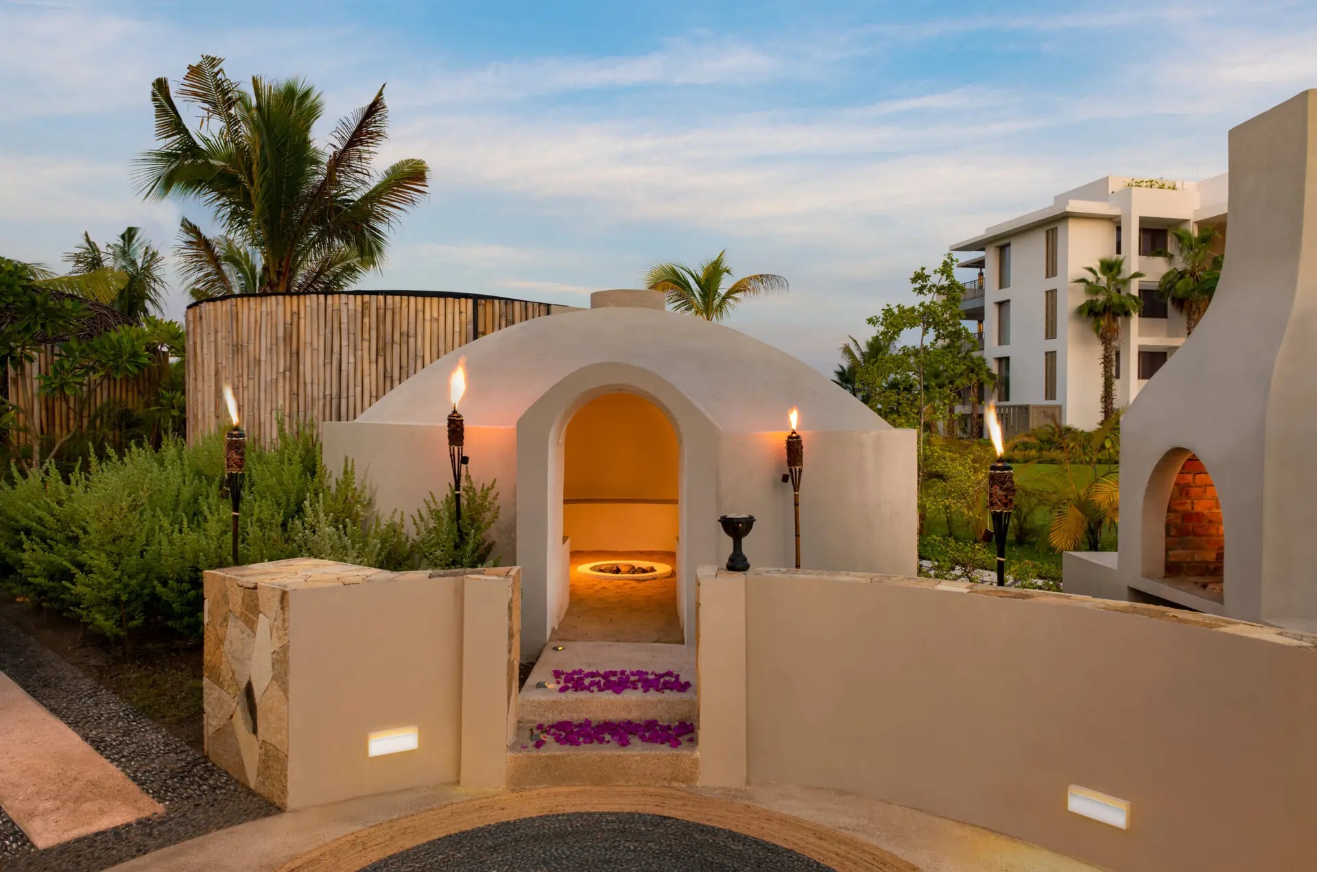 Circular clay dome structure with burning torches outside, flower petals on steps, and tropical landscaping nearby.