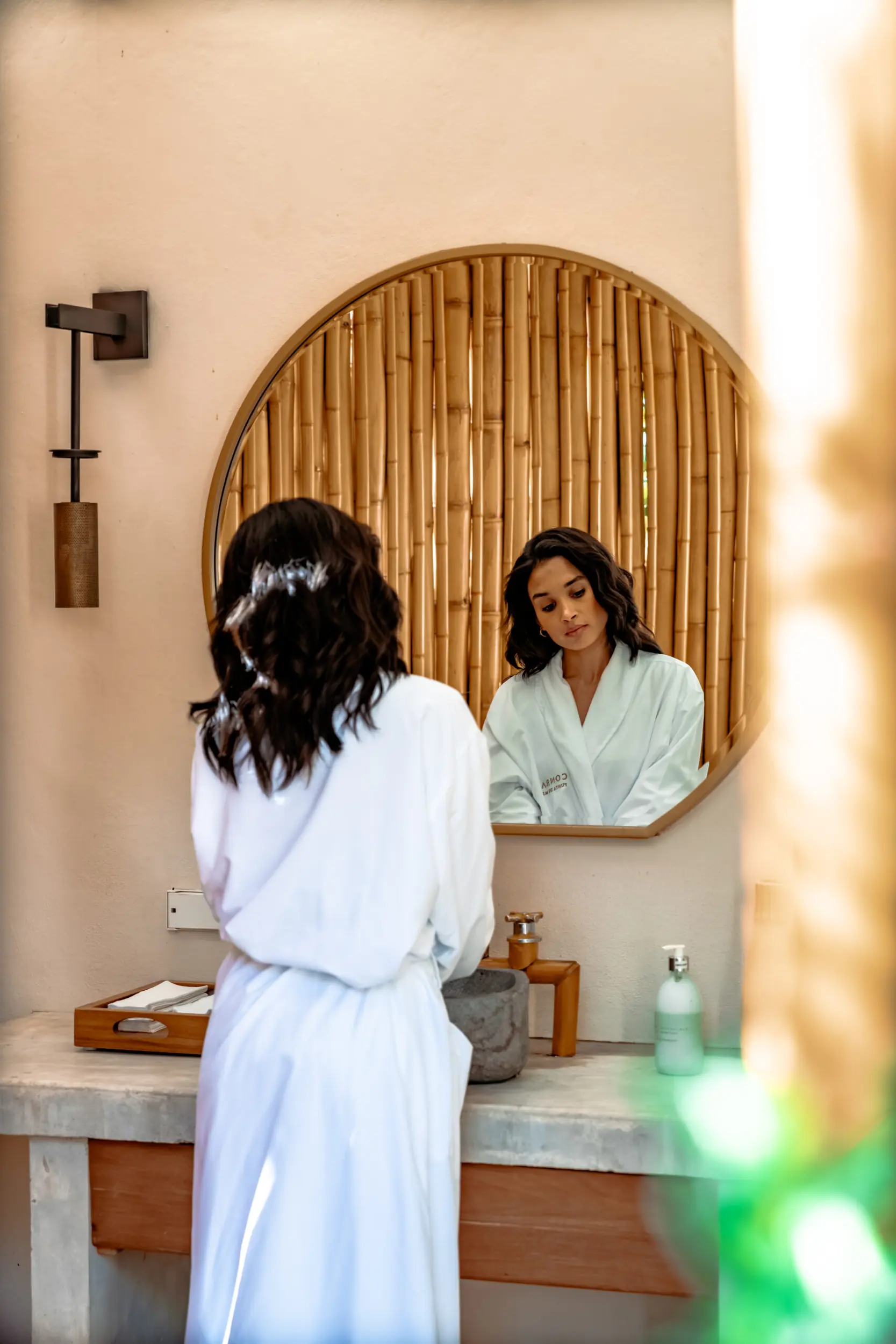 Woman in white robe stands at modern bathroom sink, reflected in round mirror with bamboo backdrop.