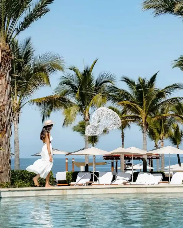 Una mujer con vestido blanco y sombrero camina junto a una piscina tropical, rodeada de palmeras.