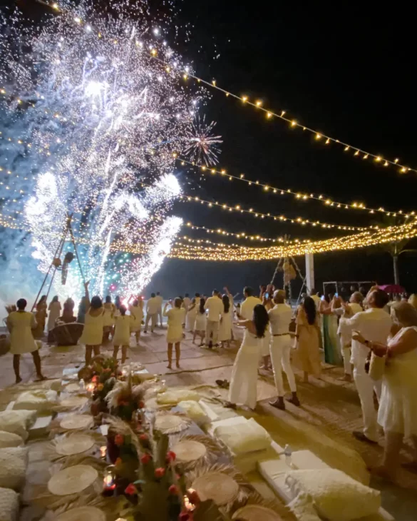 Nighttime beach party with string lights, fireworks, and guests in white gathered around low candlelit tables.