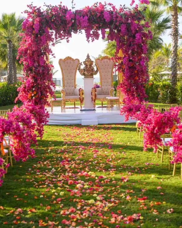 Hindu wedding ceremony setup with Ganesha statue, pink floral arch, gold chairs, and petal-lined aisle in tropical garden.
