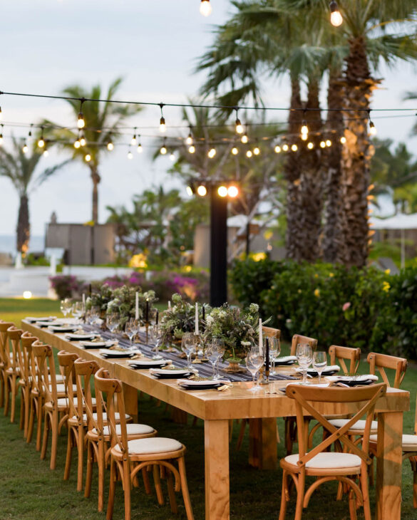 Elegant outdoor table with black plates, white cushions, floral centerpieces, and string lights in a tropical garden setting.