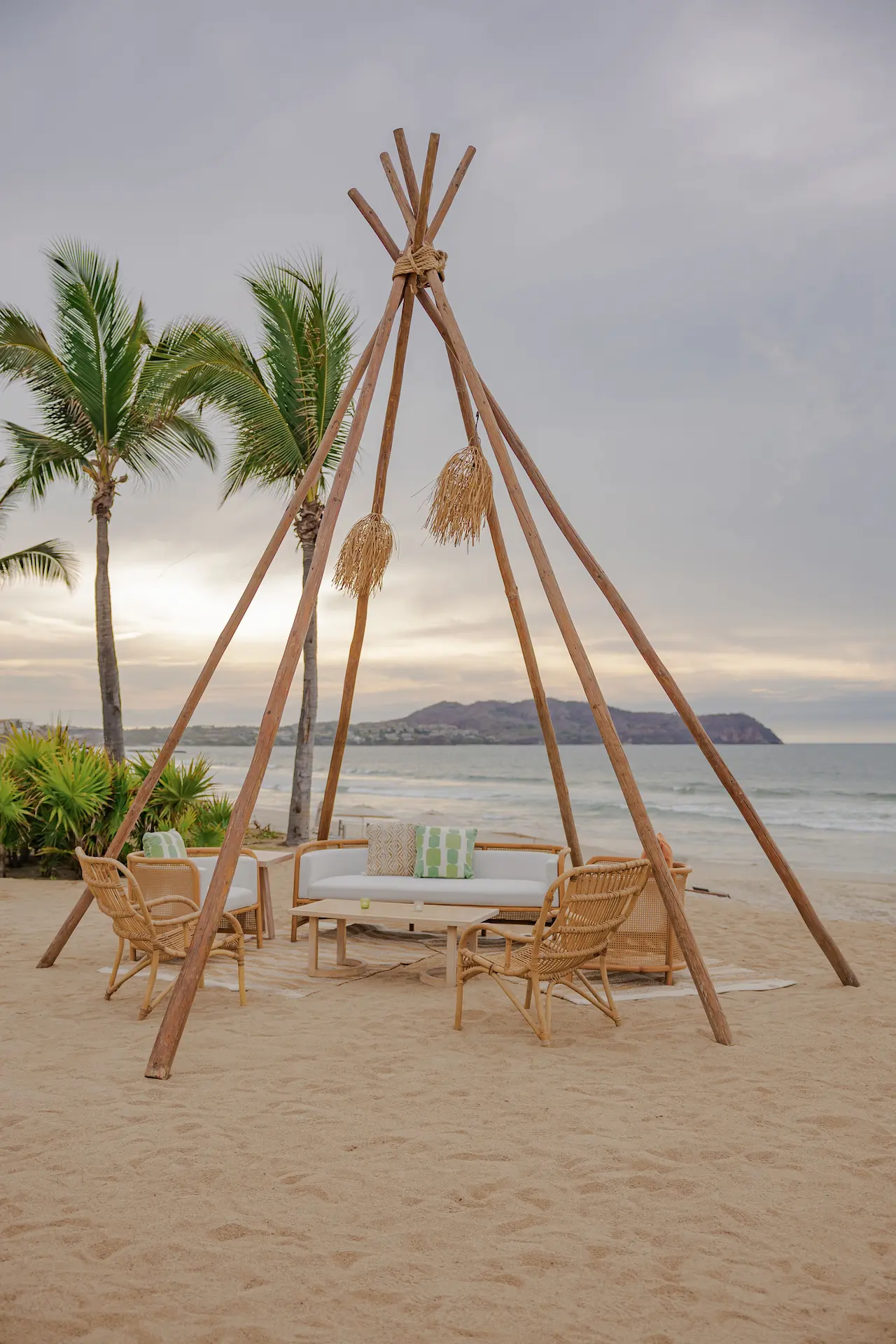 Wooden teepee structure on beach surrounds cozy seating area with chairs, sofa, table, and tropical background.