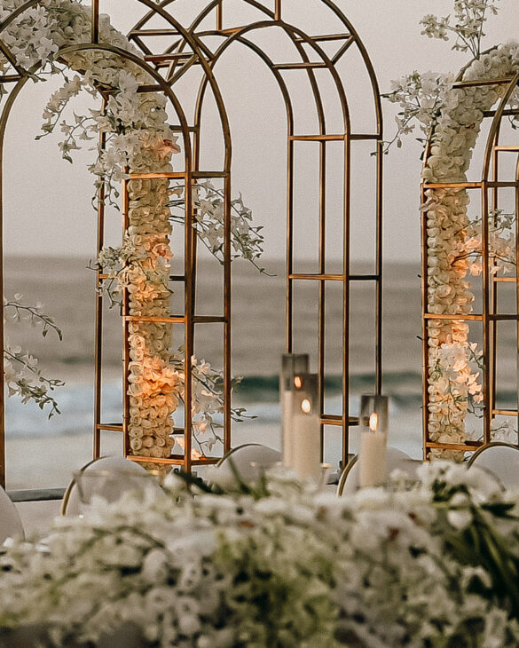 Oceanfront wedding table with white florals, gold arches, and candlelit elegance against a serene coastal backdrop.