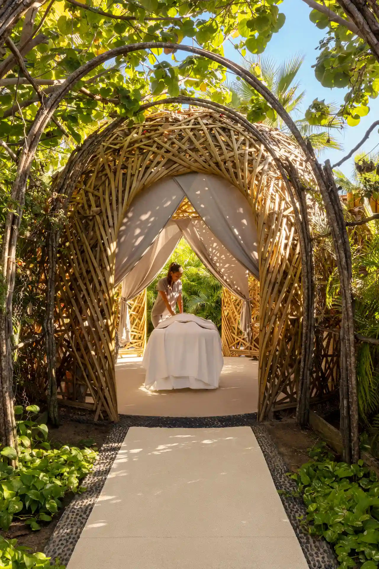 Masajista realizando un tratamiento dentro de una cabaña de madera tejida, rodeada de exuberante vegetación y luz solar filtrada.
