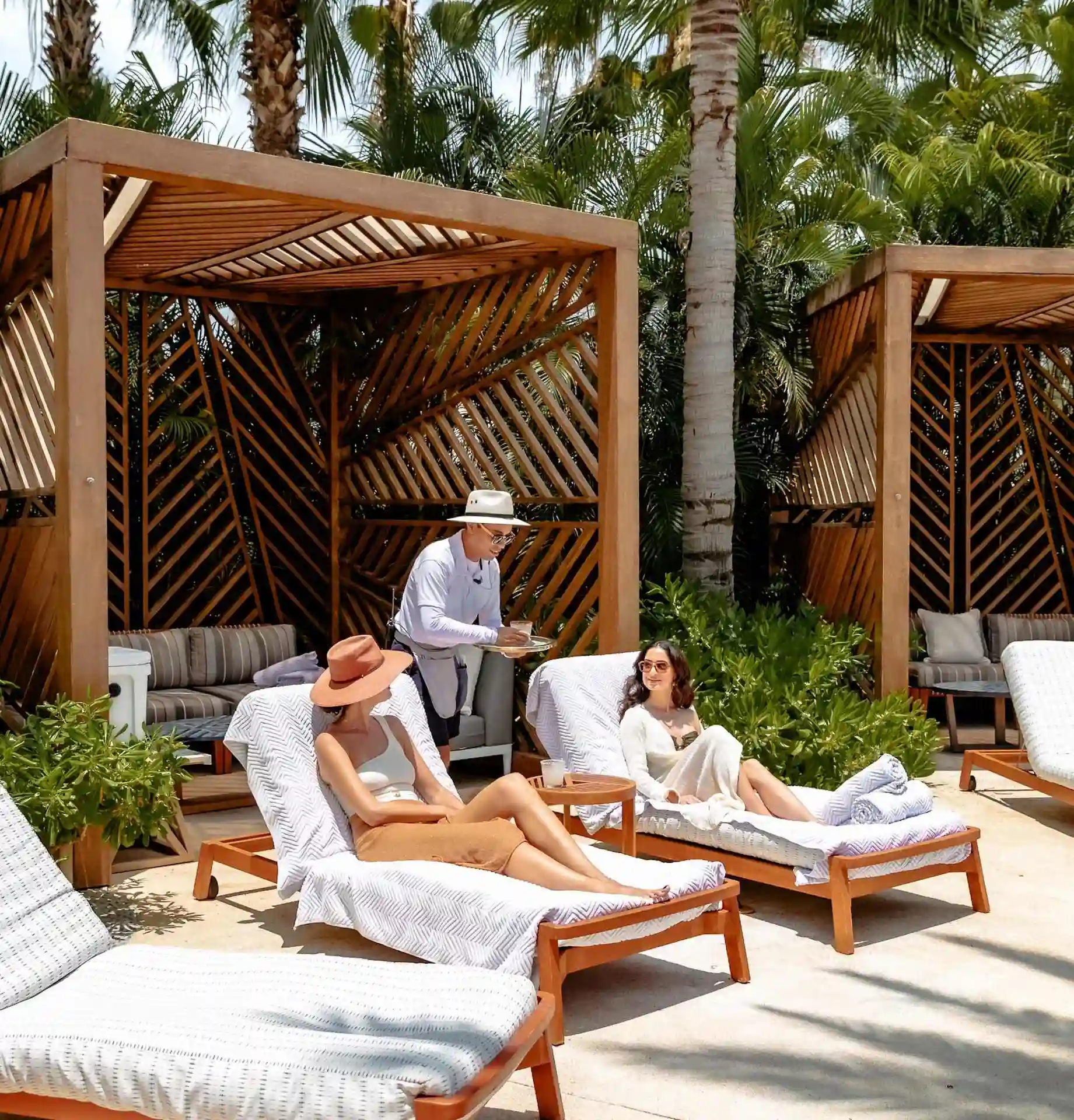 Two women relax on sun loungers by private cabanas, poolside, as a server offers refreshing drinks.