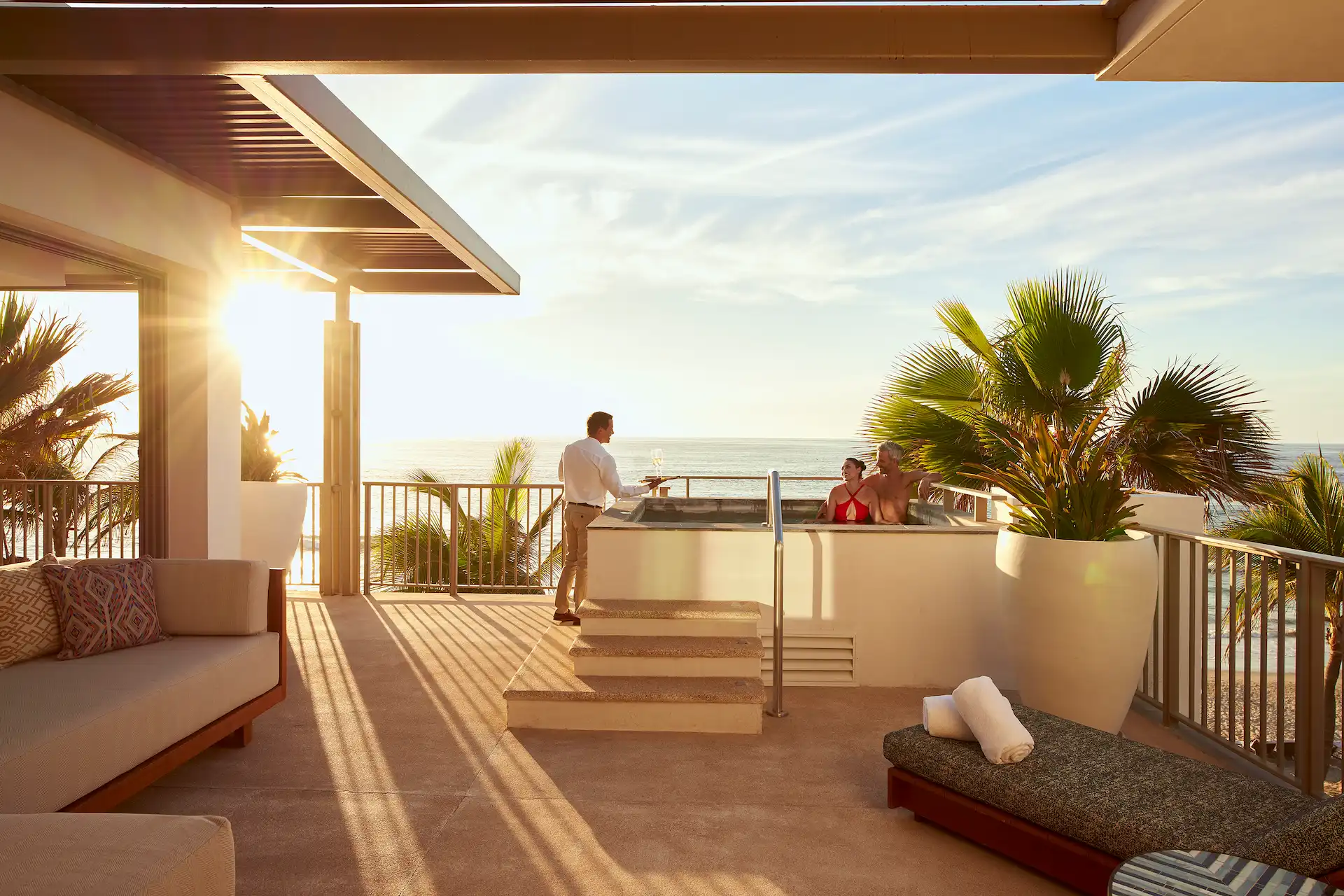 Couple relaxing in rooftop hot tub at sunset while server brings champagne, overlooking ocean.