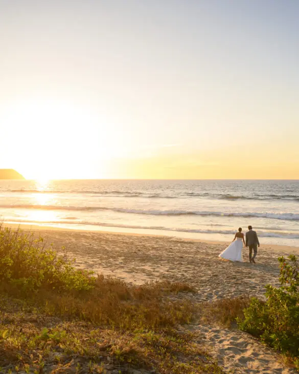 Couple walking hand-in-hand along beach at sunset with golden sky and ocean waves in the background.