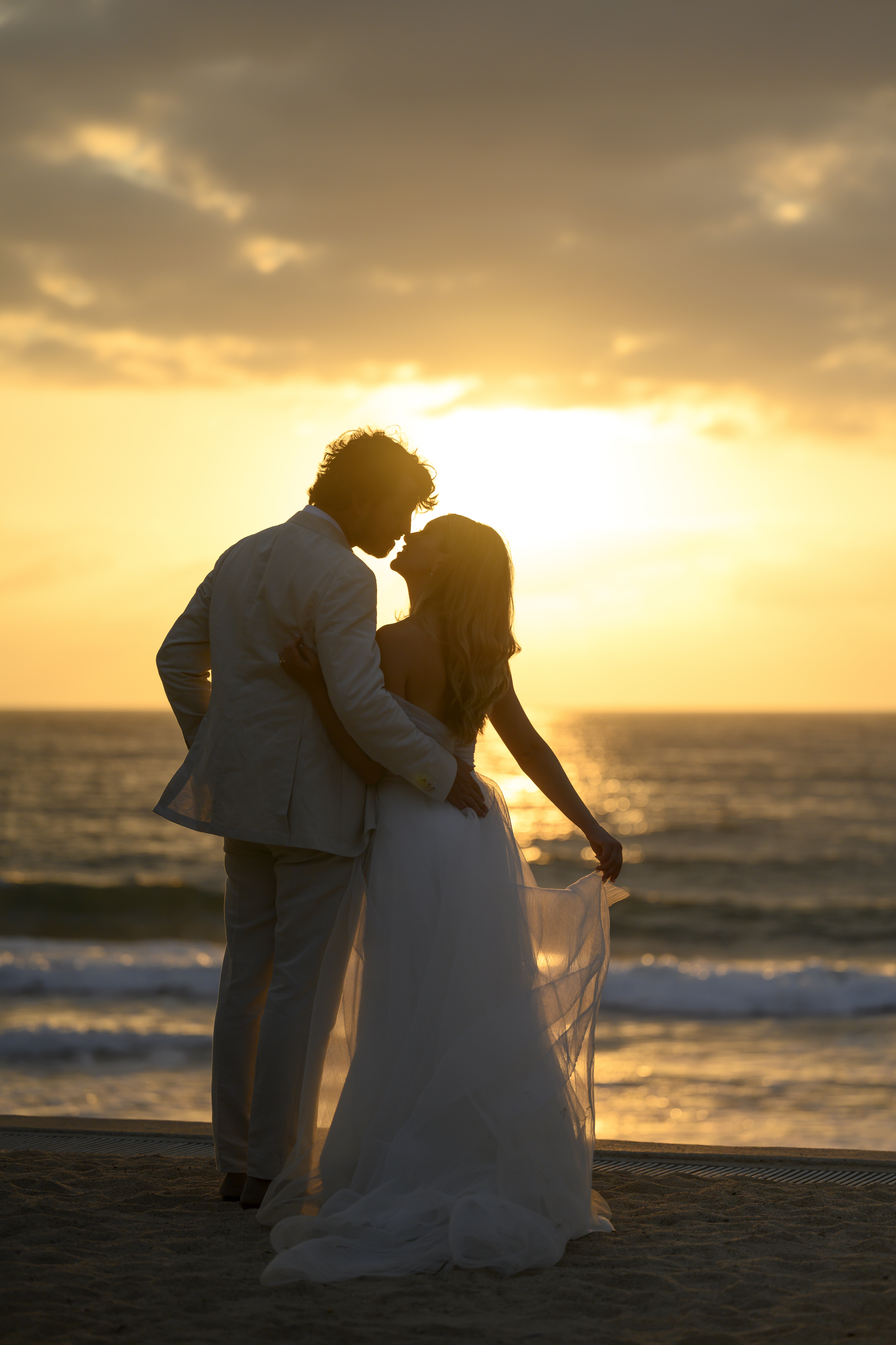 Bride and groom silhouetted kissing on the beach at sunset, waves glowing behind them.